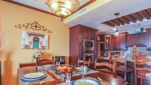 Un comedor con una mesa de madera dispuesta para cuatro personas, rodeada de sillas de madera oscura. Se aprecia una decoración en la pared sobre la mesa y un ventilador de techo.