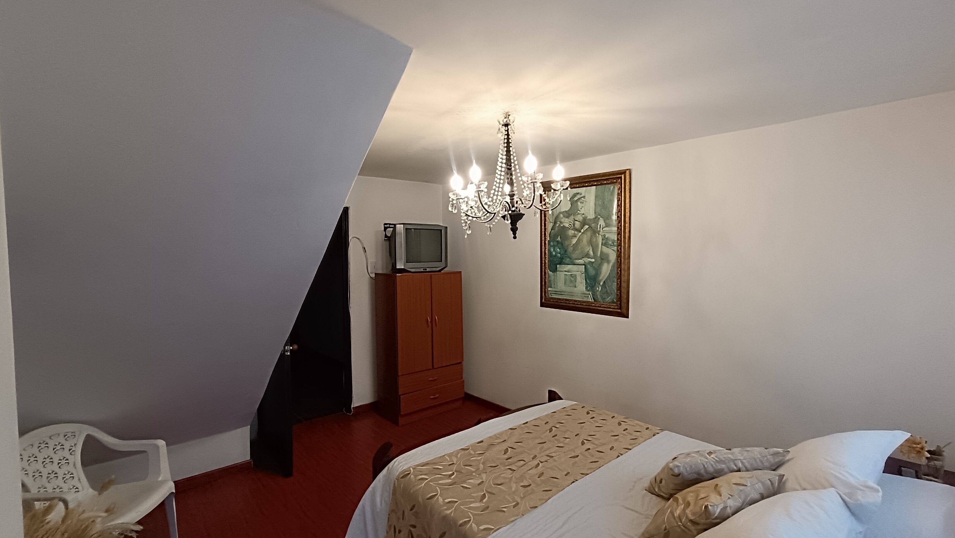 A bedroom with a bed, a television on a wooden cabinet, a chandelier, and a painting on the wall.