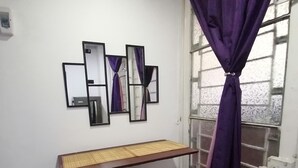 Um quarto pequeno com uma escrivaninha e dois bancos, uma janela com cortinas roxas e um espelho fixado na parede.