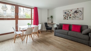 Una sala moderna con un sofá gris, cojines rojos, una mesa de comedor con sillas y un gran ventanal con cortina roja.