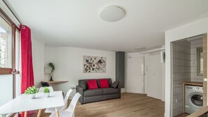 Un apartamento moderno con comedor, un sofá gris con cojines rojos, lavadora y un ventanal con cortinas rojas.