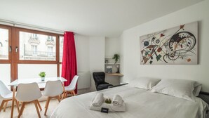 Habitación de hotel con cama, escritorio y sillas, cortina roja, un cuadro en la pared y vista a edificios exteriores.