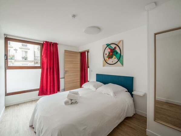 A hotel room with a bed, a window with red curtains, a wooden door, and a wall art piece.