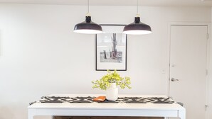 A white dining table with a black and white patterned runner, a vase with yellow flowers, and a wooden bench. Two black pendant lights hang above.