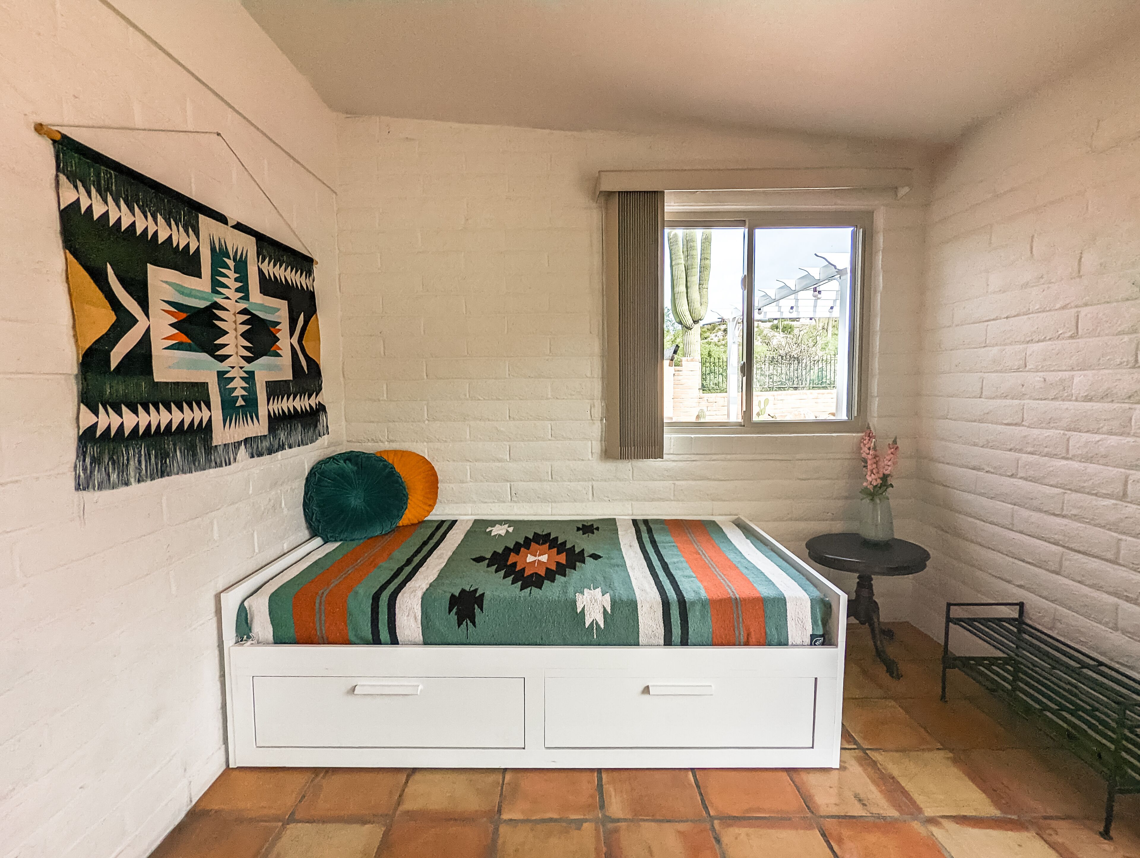 A room with a bed, a window, a wall with a geometric tapestry, and a small table with a vase.