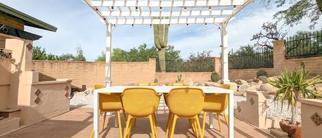Un patio exterior con una pérgola, una mesa y sillas amarillas.