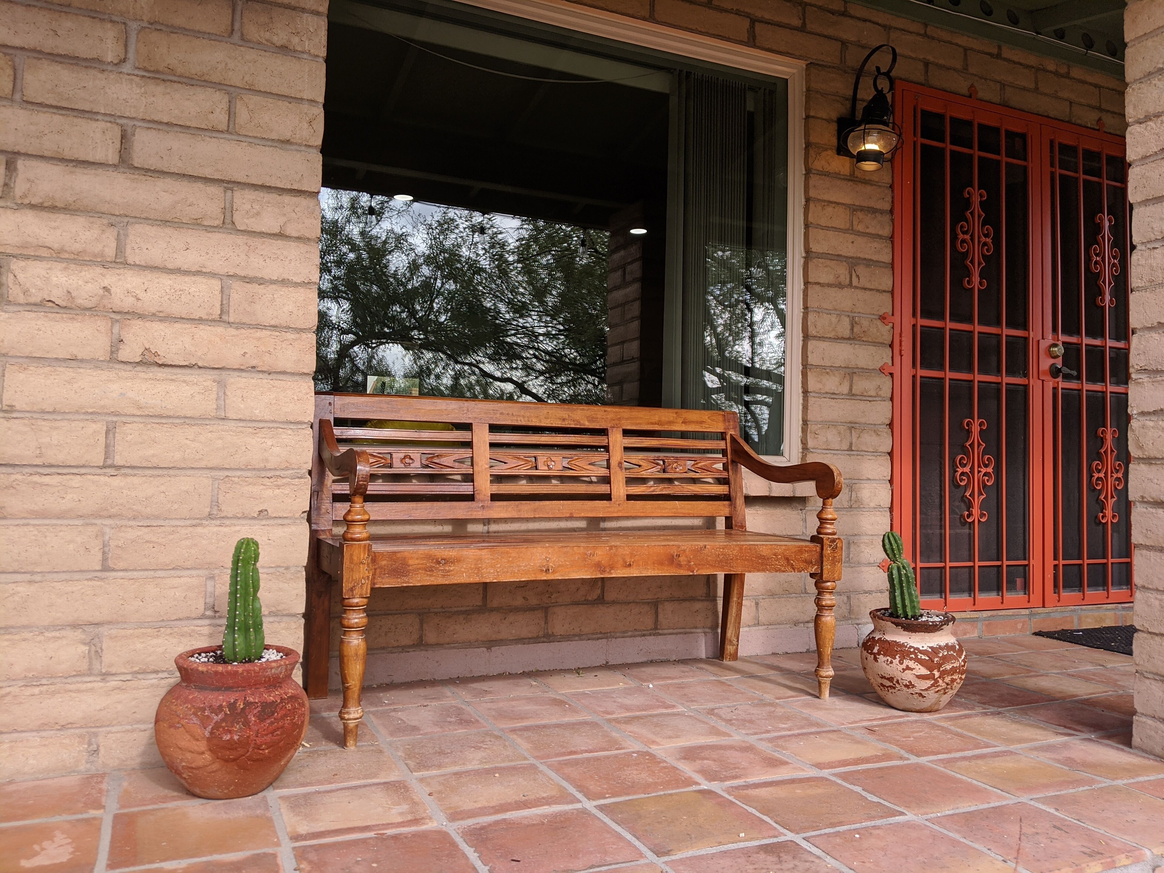 Un banc en bois doté d’un dossier décoratif, placé sur une terrasse carrelée, avec deux cactus en pot.