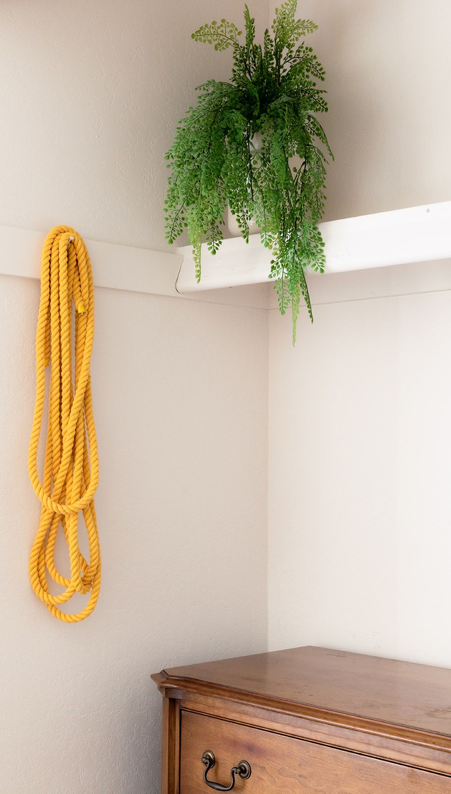 Esquina de una habitación con un aparador de madera, una cuerda amarilla y una planta verde sobre un estante blanco.