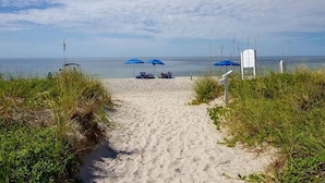 Un sentier de sable qui longe la plage et mène à l’océan, avec des parasols bleus et un panneau indicateur.