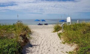 Ein sandiger Küstenpfad führt zum Meer, mit blauen Sonnenschirmen und einem Wegweiser.
