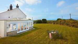 A white house with a glass conservatory, a lawn, and a table with chairs. - A beautiful, dog-friendly holiday home with magnificent views of two coastlines (Praa Sands)
