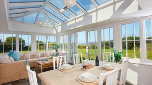 A bright, spacious conservatory with a high ceiling, large windows, and a dining area set up for a meal. - A beautiful, dog-friendly holiday home with magnificent views of two coastlines (Praa Sands)