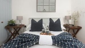 Una cama bien hecha con una colcha de diseño azul y blanco, dos almohadas, una mesita de noche con una lámpara, una bandeja con frutas y una taza, y un espejo en la pared.