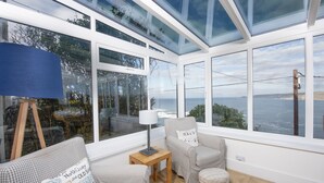 A conservatory with a view of the sea, furnished with a sofa, armchair, and a small table. - This property combines a breath-taking coastal location with modern styling (Sennen Cove)