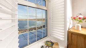 A room with a large window overlooking a beach, a wooden cabinet, and a vase with flowers. - This property combines a breath-taking coastal location with modern styling (Sennen Cove)