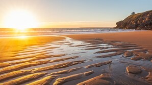 A beach with a sunset, waves, and a rocky cliff with a structure on top. - This property combines a breath-taking coastal location with modern styling (Sennen Cove)