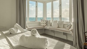 Habitació amb un gran finestral que dona al mar, un llit amb tovalloles arremolinades i una banca amb coixins.