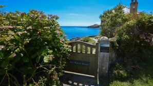 Una propietat costanera anomenada 'The Watch House' amb una porta de pedra, vegetació exuberant i una vista clara del mar.