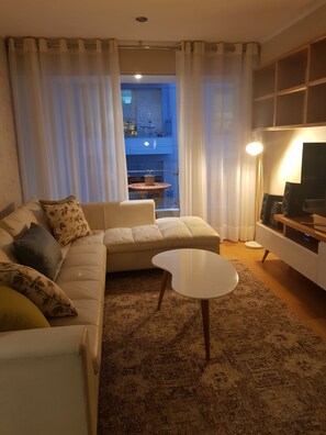 Un salon avec un canapé, une table basse et un meuble pour la télévision. On aperçoit un balcon avec une table et des chaises à travers la fenêtre.