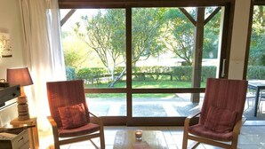 Una habitación con dos sillones rojos, una mesa de madera y un banco. Se ve un ventanal con vistas a zonas verdes.