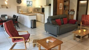 Una sala de estar acogedora con un sofá gris, cojines rojos, una mesa de centro de madera y un sillón rojo. Se ve una cocina con armarios blancos y una ventana al fondo.