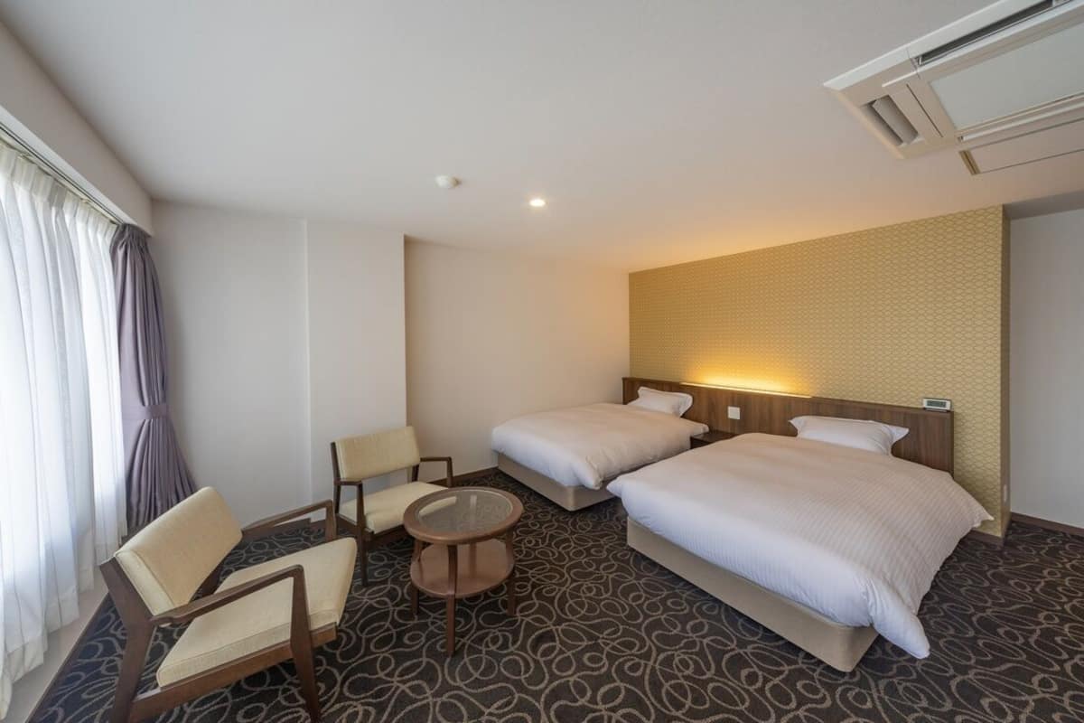 A hotel room with two beds, a chair, a small table, and a window with curtains.