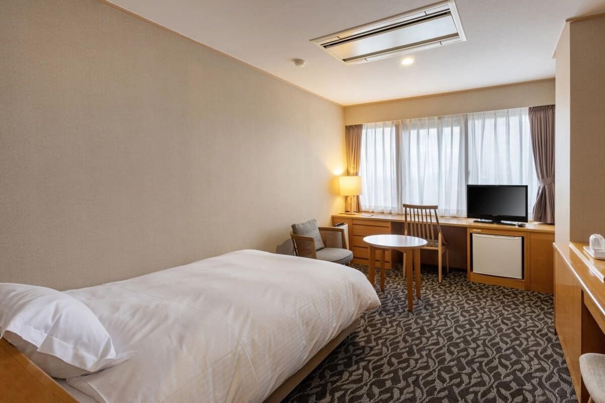 A hotel room with a bed, a desk with a chair, a television, and a window with curtains.