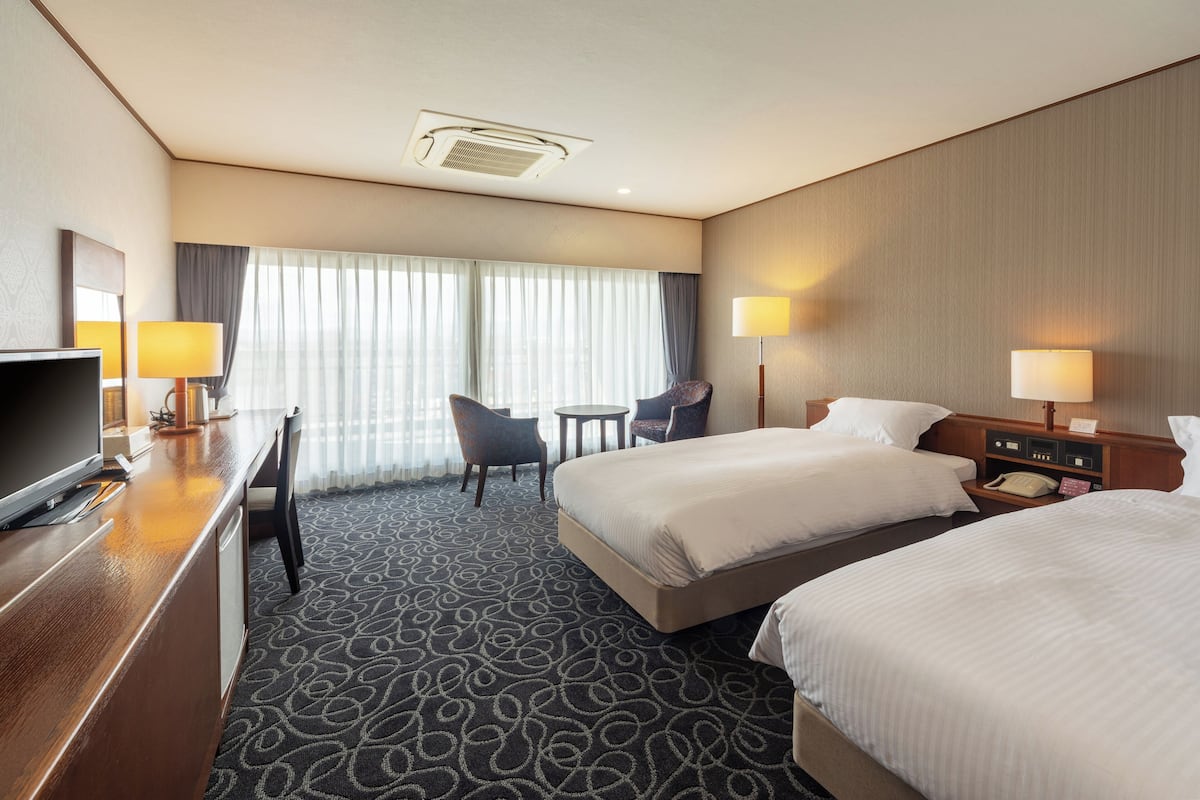 A hotel room with a bed, two bedside tables, a desk with a chair, a TV, and a window with curtains.