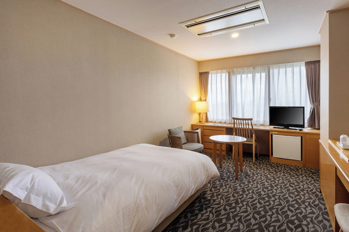 A hotel room with a bed, a desk with a television, a chair, a lamp, and a small refrigerator.