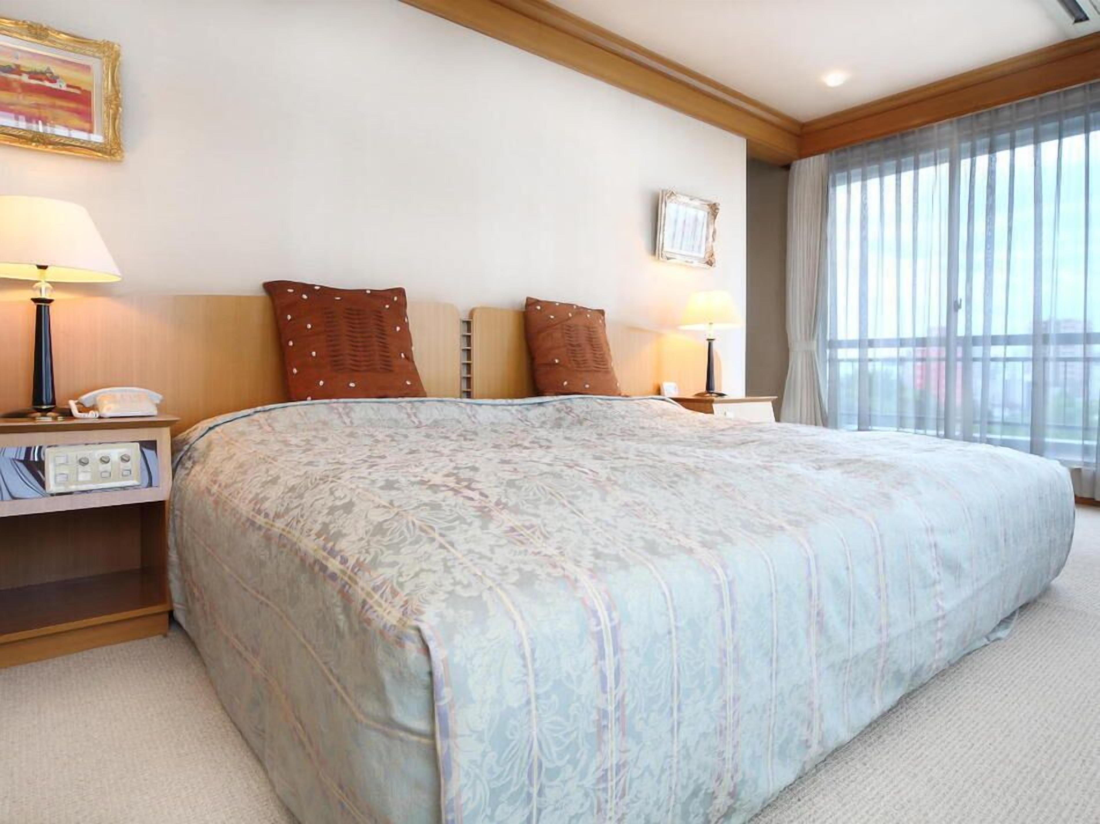 Habitación de hotel con una cama grande, lámparas de noche, una mesita de noche con teléfono, una mesita pequeña y una ventana con cortinas.
