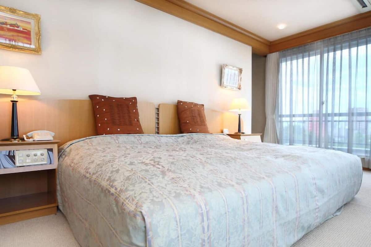 A hotel room with a large bed, bedside lamps, a nightstand with a phone, a small table, and a window with curtains.
