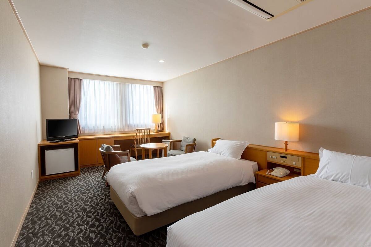 A hotel room with two beds, a desk with a television, a small table, and a chair.