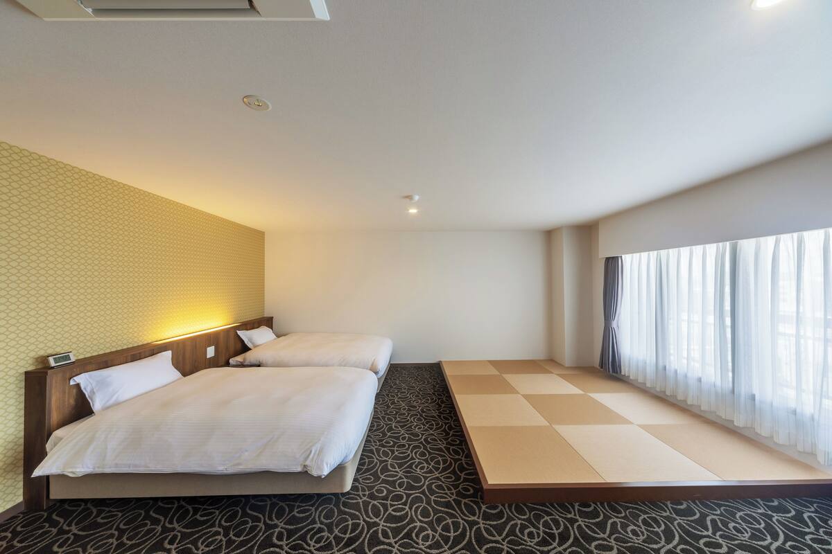 A hotel room with two beds, a tatami mat floor, a large window with curtains, and a wall with a geometric pattern.
