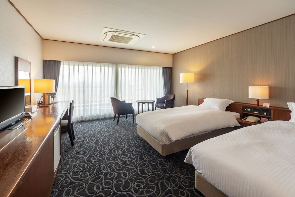 A hotel room with two beds, a desk, a chair, a TV, and a window with curtains.