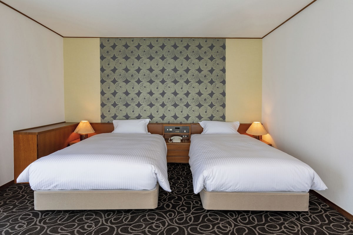 A hotel room with two beds, a wooden headboard, a patterned wall, and a small table with a lamp.
