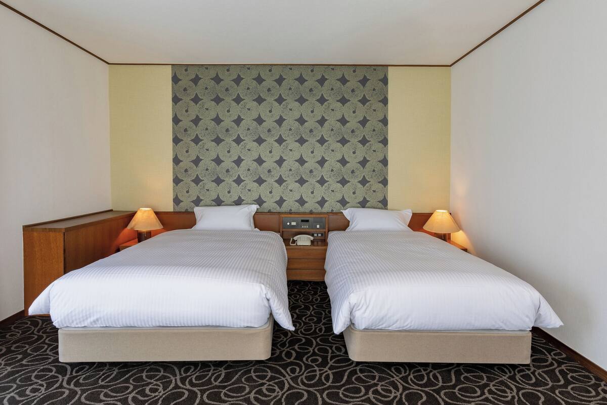 A hotel room with two beds, a wooden headboard, a patterned wall, and a small table with a lamp.