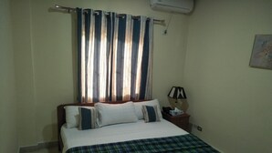 A bedroom with a bed, pillows, a nightstand, a lamp, and a window with curtains. - Diana Hotel (Monrovia)