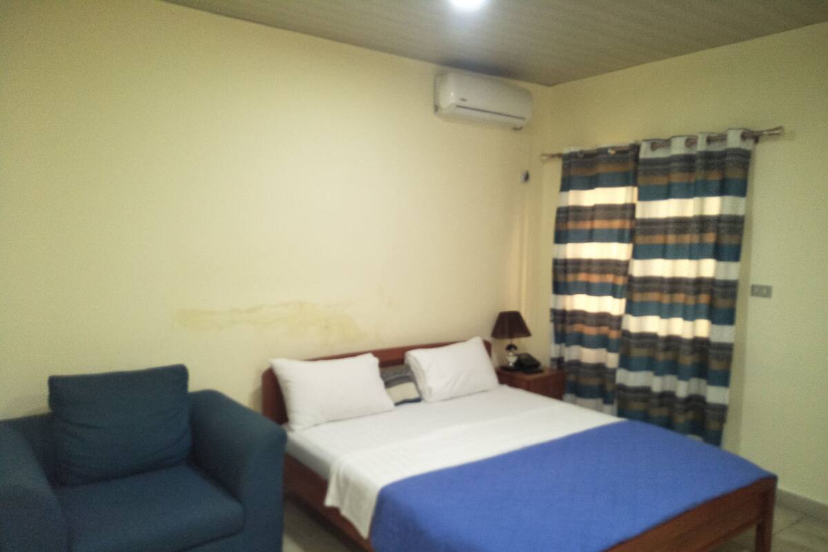A bedroom with a bed, a chair, a nightstand, an air conditioner, and a curtain.