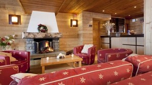 A cozy log cabin interior with a fireplace, red upholstered furniture, and a kitchen area.