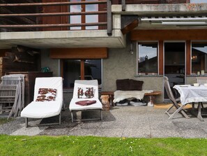 A rustic outdoor seating area with lounge chairs and a wooden table, set against a building with a balcony and large windows.