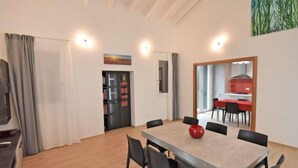 Una sala da pranzo moderna con un tavolo grande, sedie e un vaso rosso. Si intravede una cucina attraverso una porta aperta, e una libreria con libri.