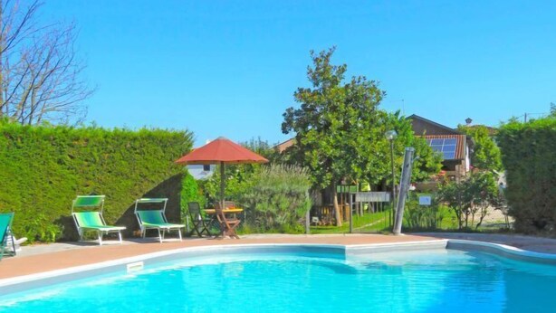 Una piscina con lettini e un ombrellone rosso, circondata dal verde e con una casa sullo sfondo.