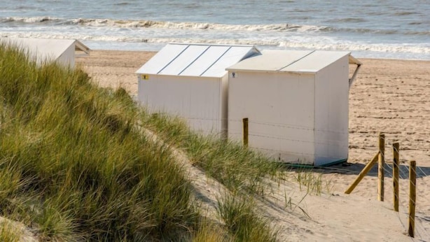 Une plage avec deux cabanes de plage blanches, un rivage sablonneux et un voilier à l’horizon.