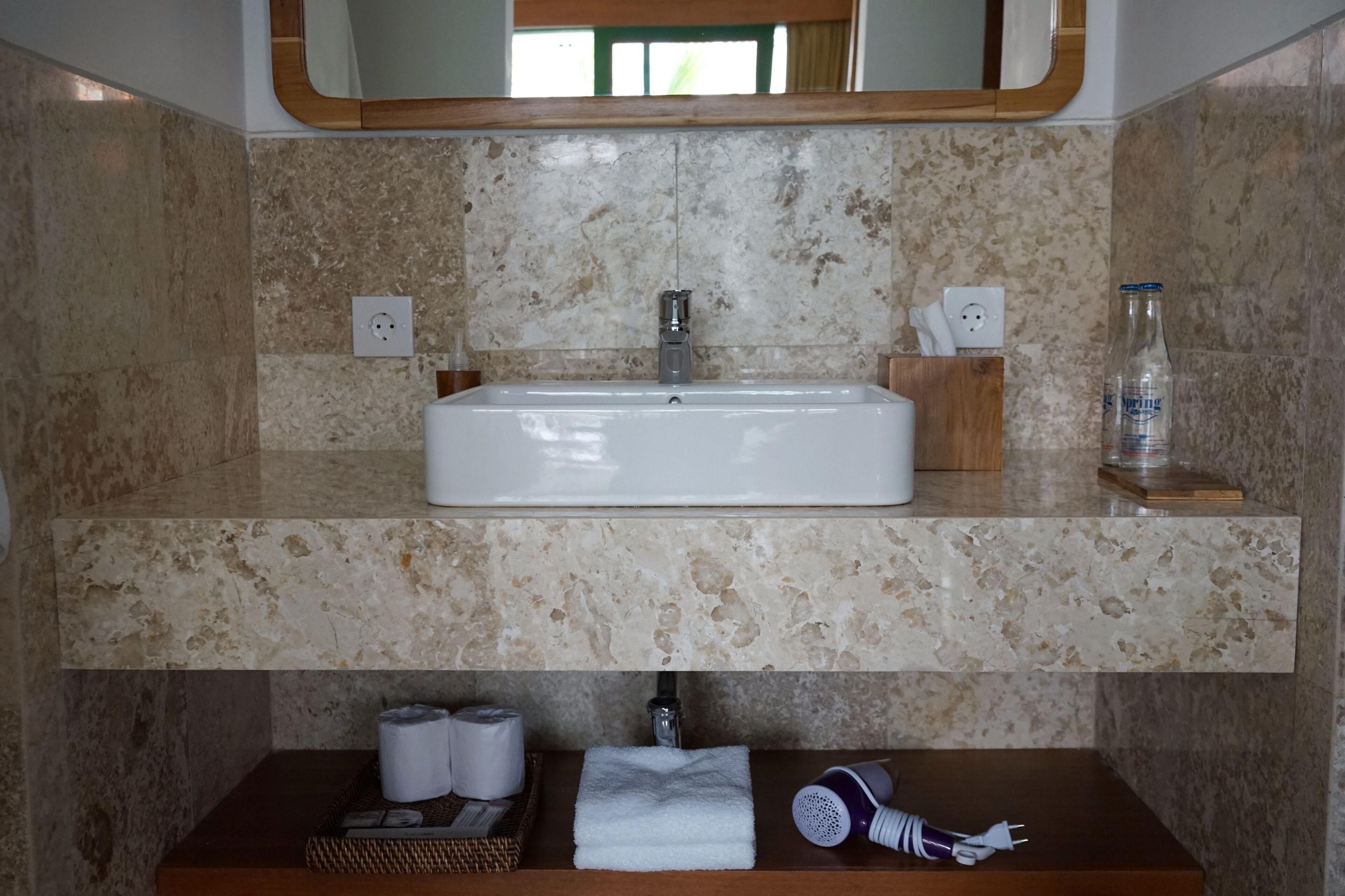 Un baño con un lavabo rectangular, paredes de mármol, un espejo sobre el lavabo y un estante inferior con artículos de aseo.