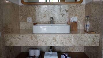 Un baño con un lavabo rectangular, paredes de mármol, un espejo sobre el lavabo y un estante inferior con artículos de aseo.