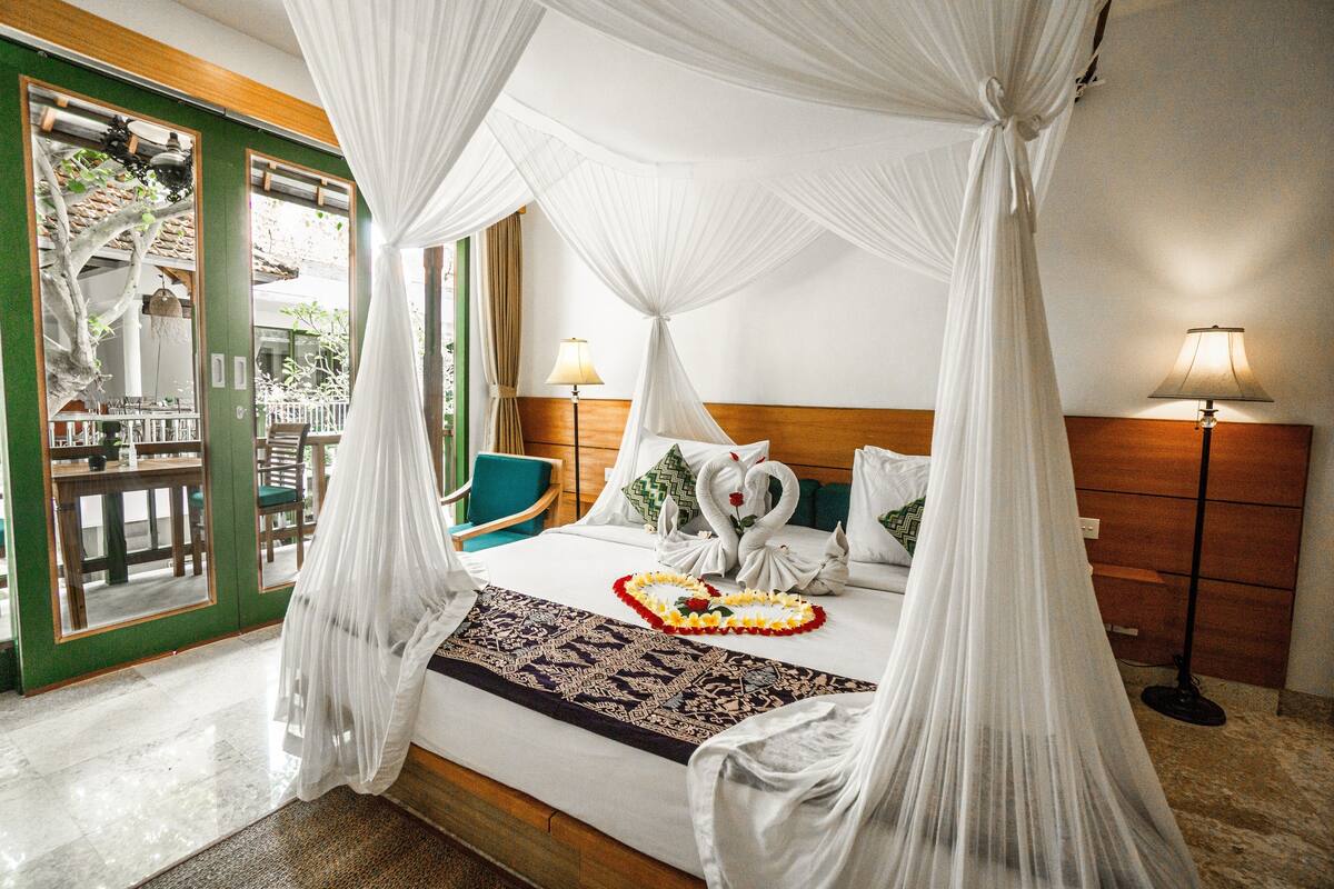 Habitación de hotel con una cama, cabecera de madera, mesita de noche con lámpara, vistas al exterior y cortinas.