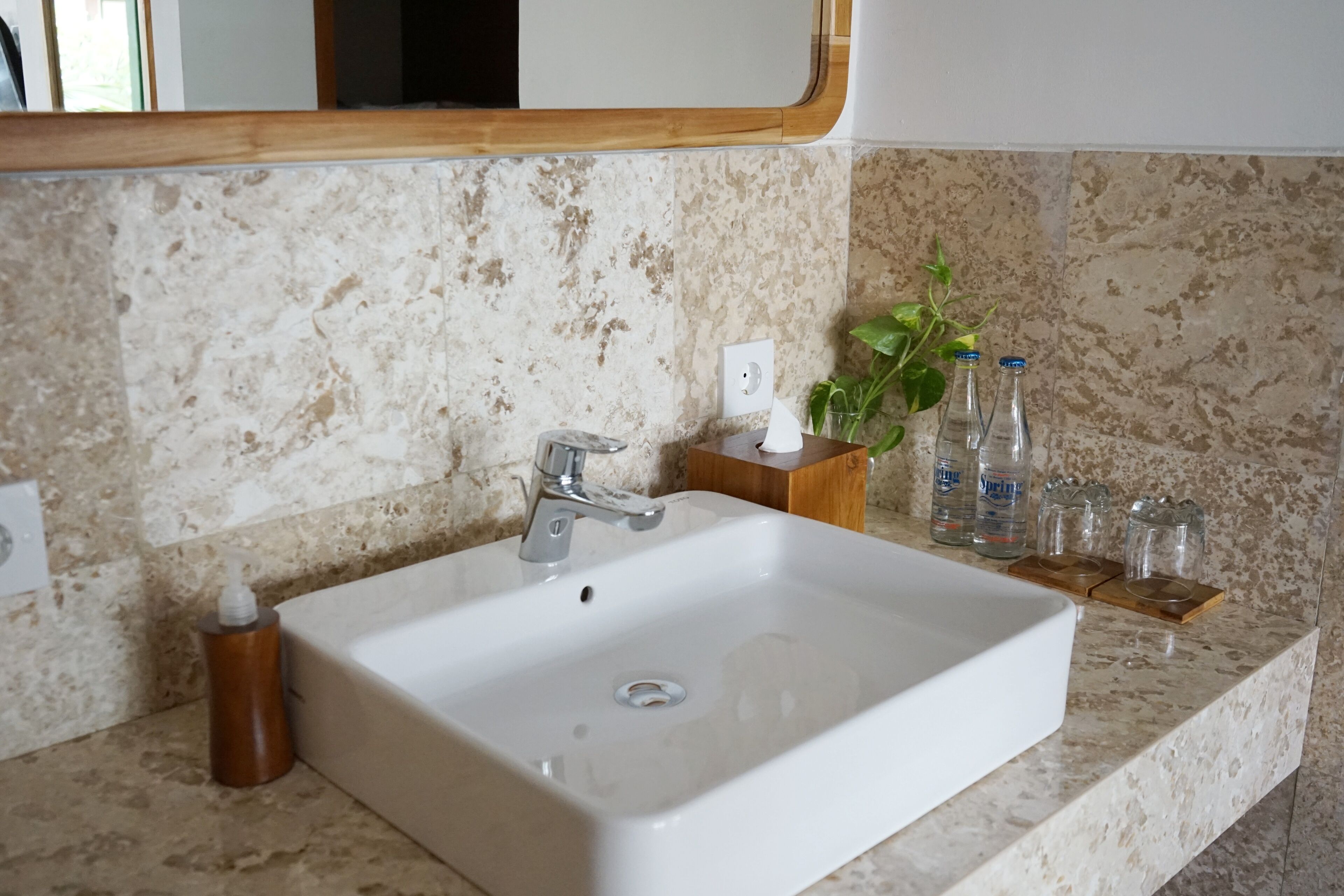 Une salle de bain avec un lavabo blanc, un distributeur de savon en bois et une étagère contenant des bouteilles et des verres.
