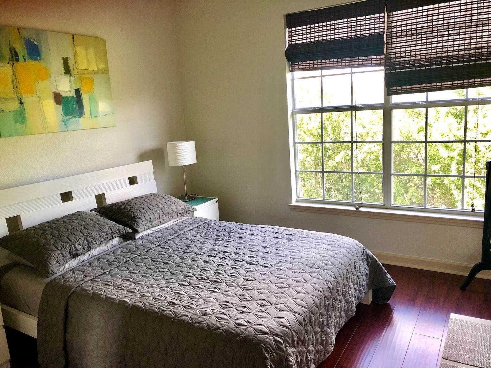 A bedroom with a large bed, a nightstand with a lamp, a window with blinds, and a painting on the wall.