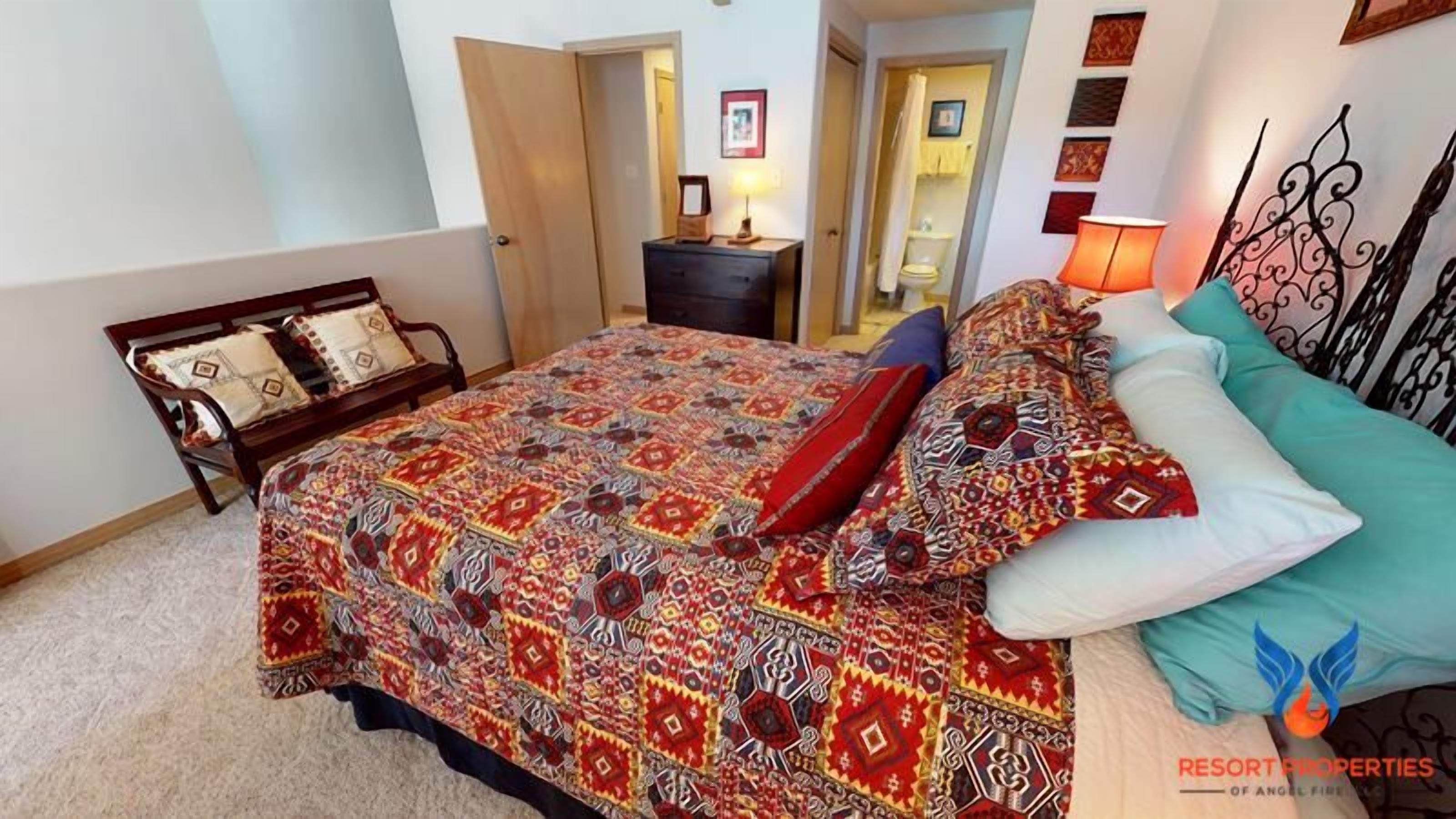 Ein Schlafzimmer mit einem großen Bett, einem Holfnachtkästchen, einem Schreibtisch und einem Stuhl. Im Hintergrund ist ein Badezimmer erkennbar.