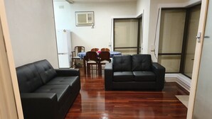 A living room with a wooden floor, two black sofas, a dining table with chairs, and a sliding glass door.